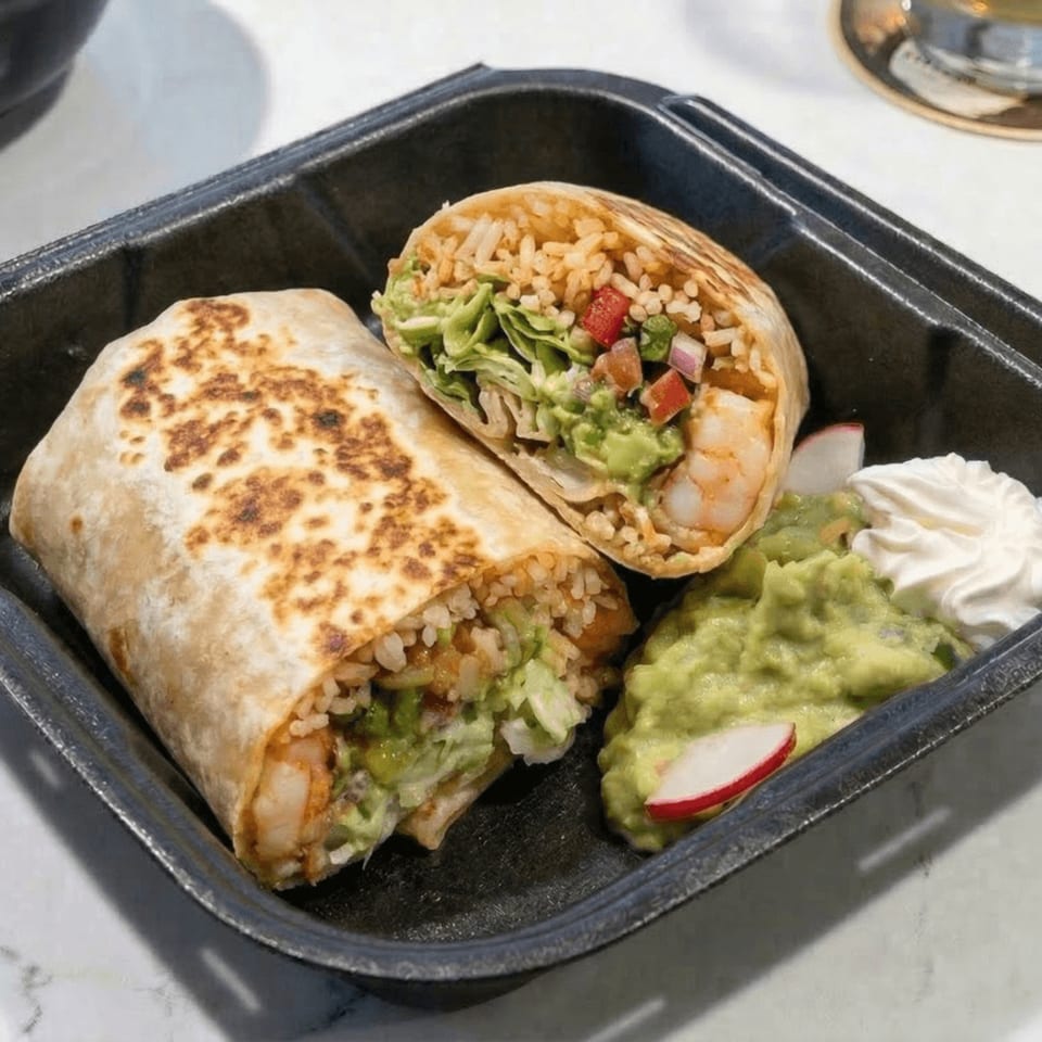 A shrimp burrito, cut in half, with guacamole and sour cream in a black container.